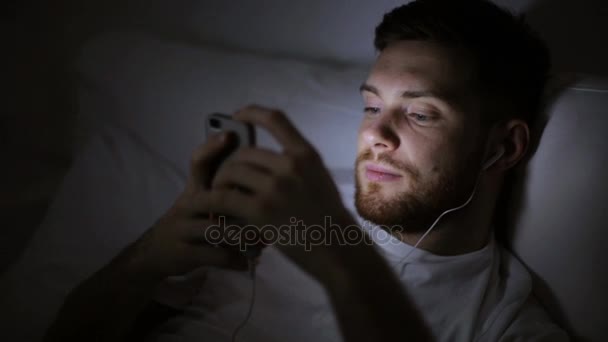 Man with smartphone and earphones in bed at night — Stock Video