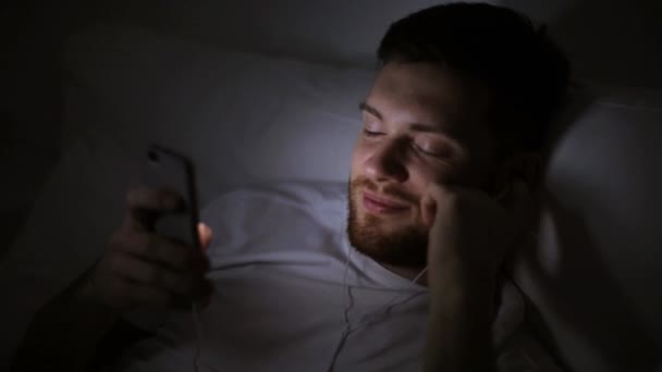 Homme avec smartphone et écouteurs au lit la nuit — Video