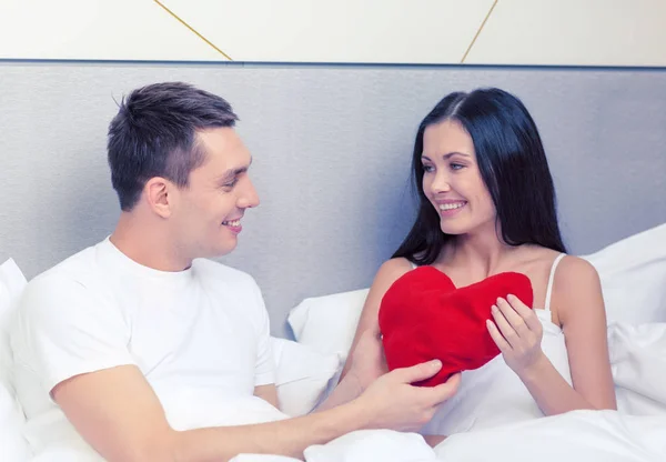 Улыбающаяся пара в постели с красной подушкой в форме сердца — стоковое фото