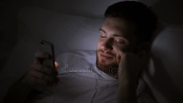 Mężczyzna ze smartfonem i słuchawkami w łóżku w nocy — Wideo stockowe