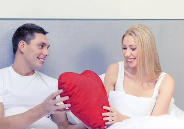 Casal sorridente na cama com travesseiro em forma de coração vermelho — Fotografia de Stock