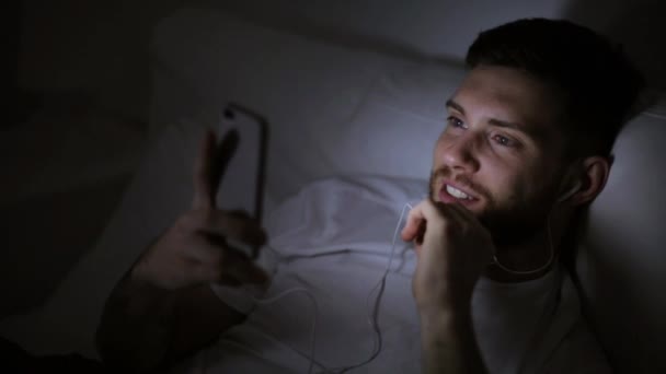 Человек со смартфоном и наушниками в постели ночью — стоковое видео