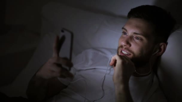 Man met smartphone en oortjes 's nachts in bed — Stockvideo