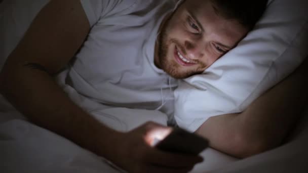 Hombre con smartphone y auriculares en la cama por la noche — Vídeos de Stock