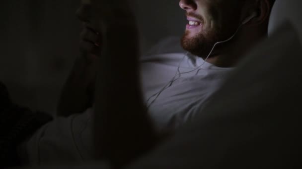 Homem com smartphone e fones de ouvido na cama à noite — Vídeo de Stock