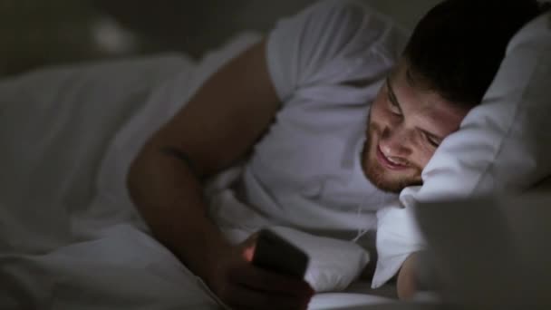 Чоловік зі смартфоном та навушниками в ліжку вночі — стокове відео