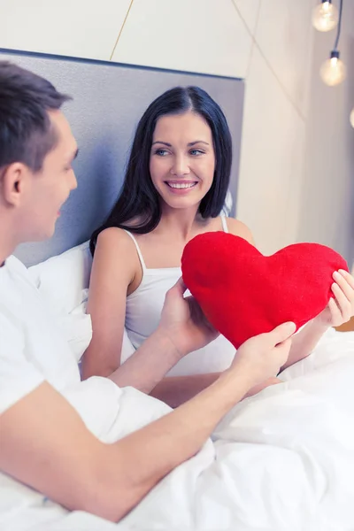 Pareja sonriente en la cama con almohada roja en forma de corazón — Foto de Stock