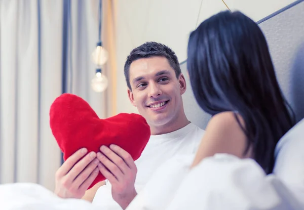 Casal sorridente na cama com travesseiro em forma de coração vermelho — Fotografia de Stock