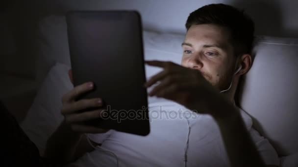 Man met tablet pc en oortelefoons in bed bij nacht — Stockvideo