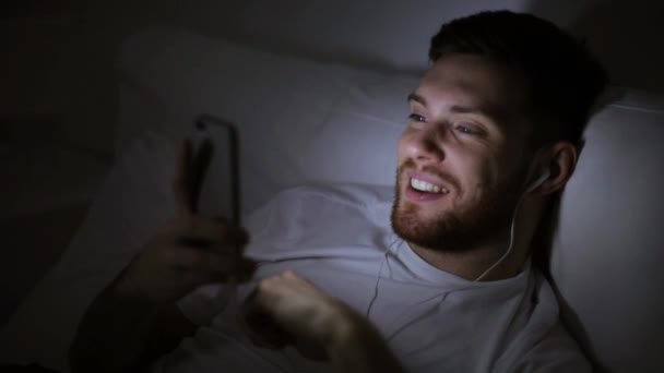 Man met smartphone en oortjes 's nachts in bed — Stockvideo