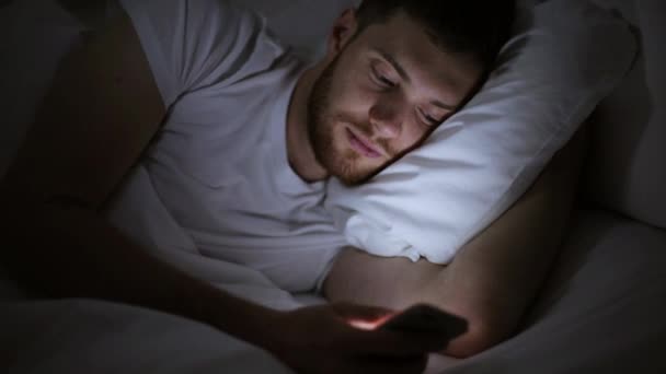 Młody człowiek ze smartfonem w łóżku w nocy — Wideo stockowe