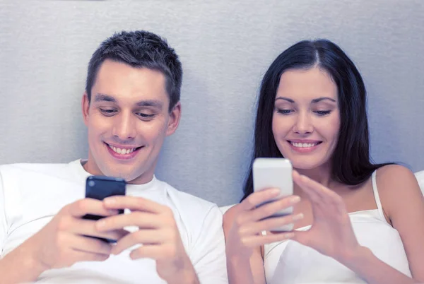 Улыбающаяся пара в постели со смартфонами — стоковое фото