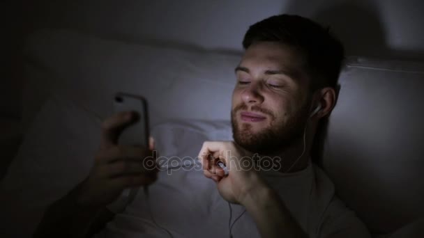Férfi okostelefonnal és fülhallgatóval az ágyban éjszaka — Stock videók