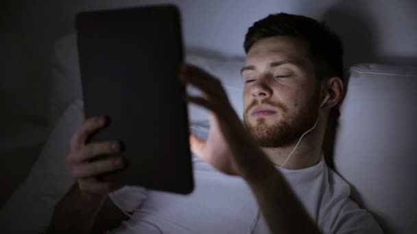 Man met tablet pc en oortelefoons in bed bij nacht — Stockvideo