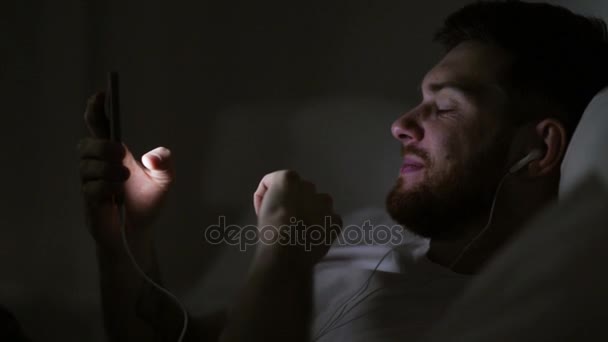 Homme avec smartphone et écouteurs au lit la nuit — Video