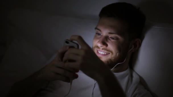 Muž s chytrým telefonem a sluchátky v posteli v noci — Stock video