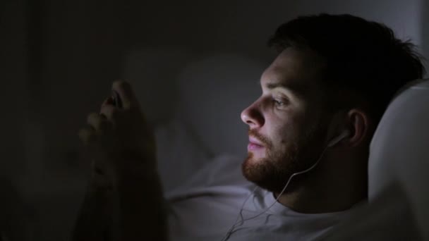 Homem com smartphone e fones de ouvido na cama à noite — Vídeo de Stock