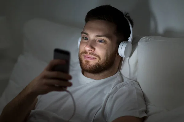 Человек со смартфоном и наушниками в постели ночью — стоковое фото