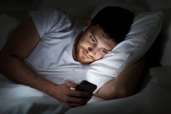 Homem com smartphone e fones de ouvido na cama à noite — Fotografia de Stock