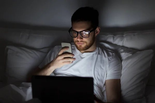 Hombre con portátil y teléfono inteligente por la noche en la cama —  Fotos de Stock