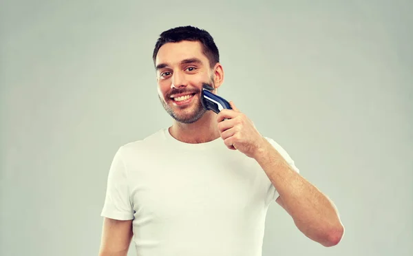 Улыбающийся мужчина бреет бороду с триммером над серой — стоковое фото