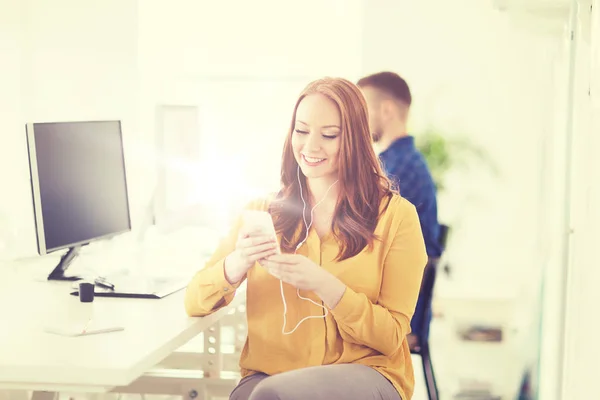 Kvinne med øretelefoner og smarttelefon ved kontoret – stockfoto