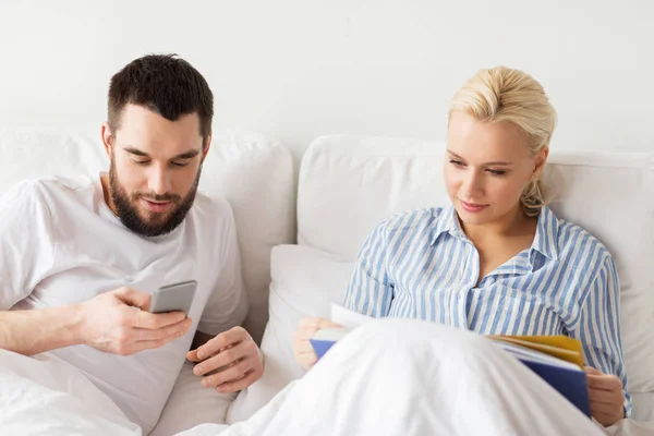 Casal com livro e smartphones na cama em casa — Fotografia de Stock