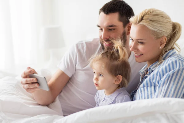 Familia feliz con teléfono inteligente en la cama en casa —  Fotos de Stock