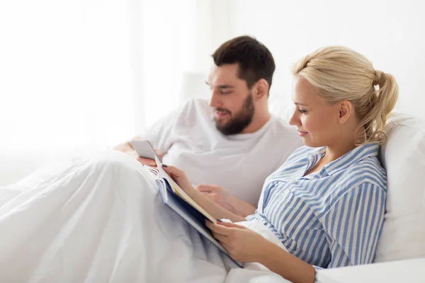 Echtpaar met boek en smartphone in bed thuis — Stockfoto