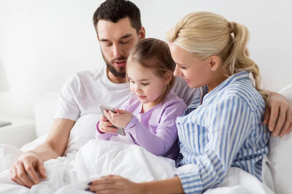 Gelukkige familie met smartphone in bed thuis — Stockfoto