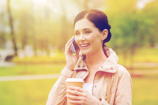 Mujer sonriente con teléfono inteligente y café en el parque —  Fotos de Stock
