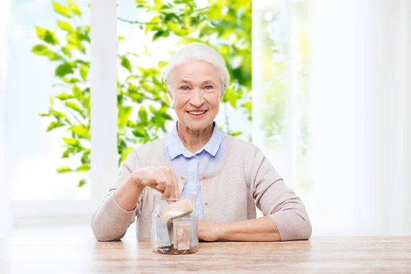 Donna anziana mettendo soldi in vaso di vetro a casa — Foto Stock