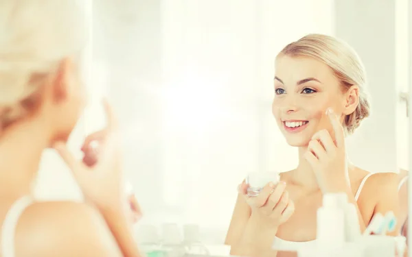Happy woman applying cream to face at bathroom — Stock Photo, Image