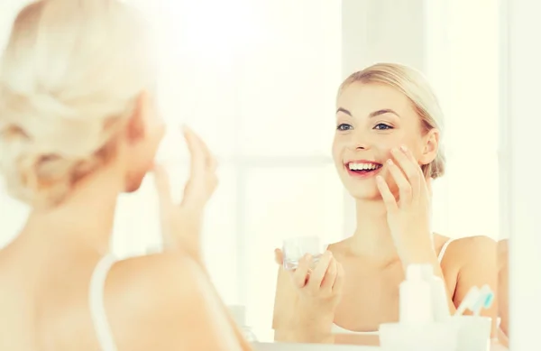 Щаслива жінка наносить крем обличчям у ванній — стокове фото