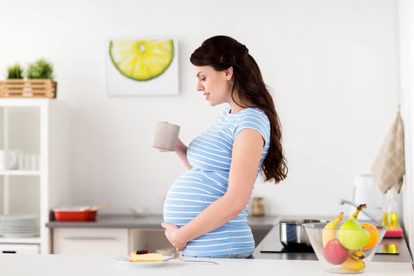 Glücklich schwangere Frau mit Becher und Kuchen zu Hause — Stockfoto