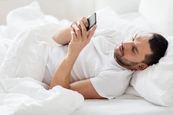 Joven con teléfono inteligente en la cama por la mañana — Foto de Stock