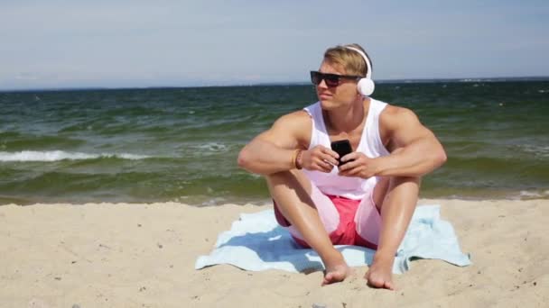 Young man with smartphone and headphones on beach — Stock Video