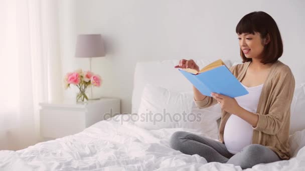 Счастливая беременная азиатка читает книгу в постели — стоковое видео