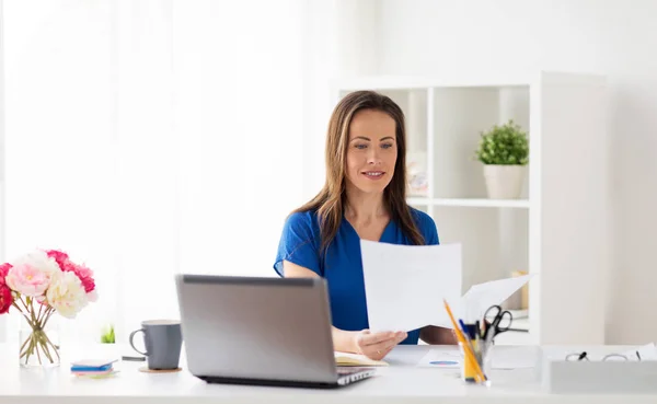 Счастливая женщина с бумагами и ноутбуком в офисе — стоковое фото