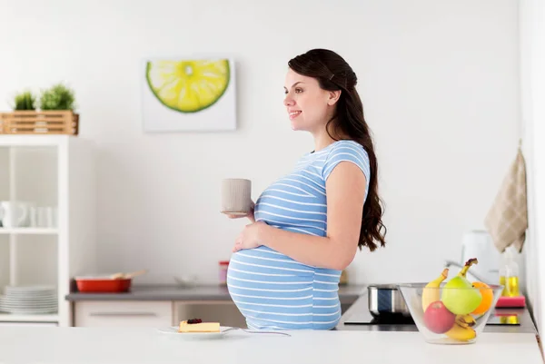 Mujer embarazada feliz con taza y pastel en casa —  Fotos de Stock