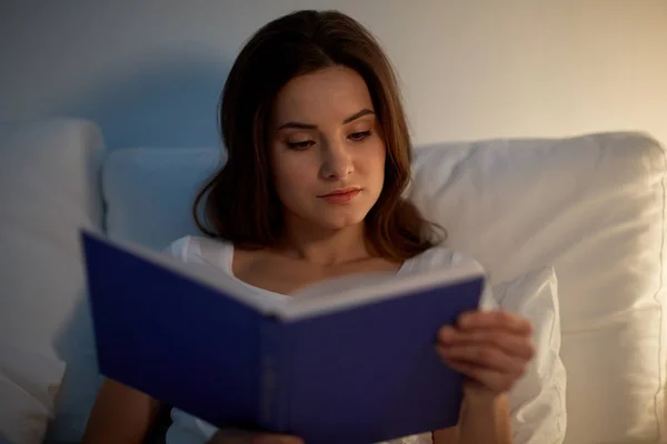 Mujer joven leyendo libro en la cama en casa de noche — Foto de Stock