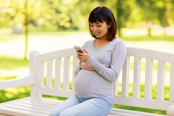 Happy těhotná Asijské žena s smartphone v parku — Stock fotografie