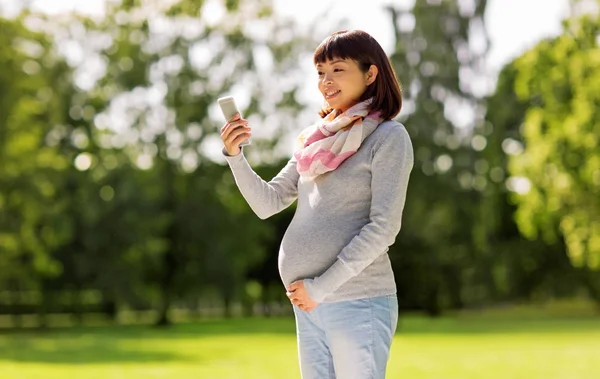 Счастливая беременная азиатка делает селфи в парке — стоковое фото
