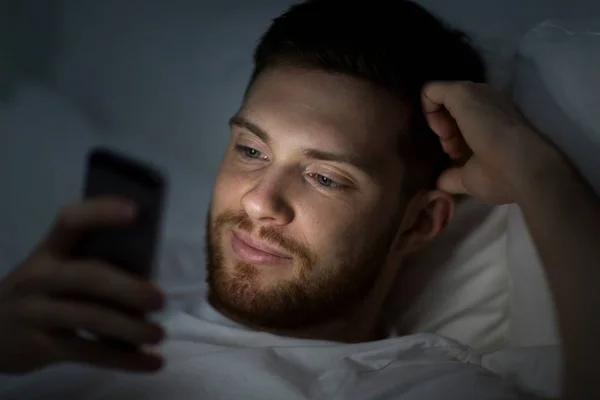 Joven feliz con teléfono inteligente en la cama por la noche —  Fotos de Stock