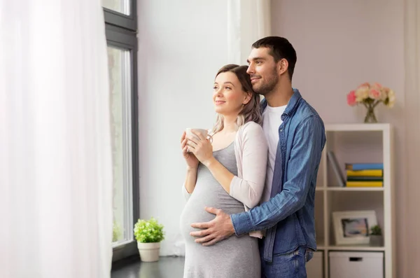 Homem abraçando mulher grávida na janela em casa — Fotografia de Stock