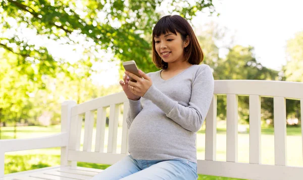 Happy těhotná Asijské žena s smartphone v parku — Stock fotografie