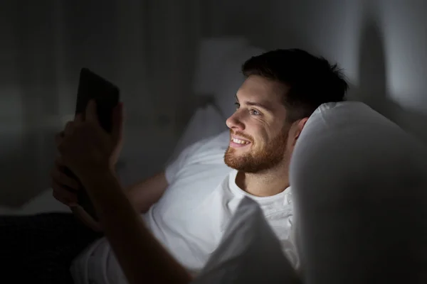 Hombre joven con tableta pc en la cama en el dormitorio en casa —  Fotos de Stock