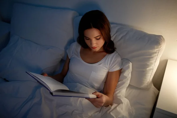 Mujer joven leyendo libro en la cama en casa de noche — Foto de Stock