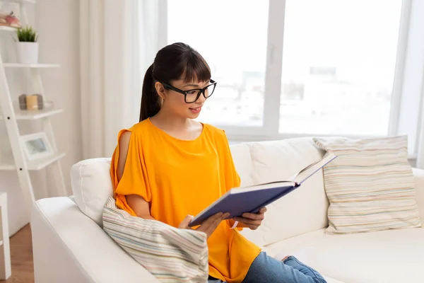 Jovem mulher asiática em óculos leitura livro em casa — Fotografia de Stock