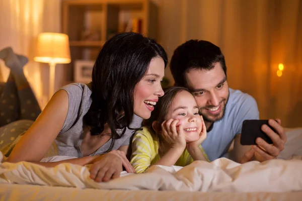 Счастливая семья со смартфоном в постели по ночам — стоковое фото